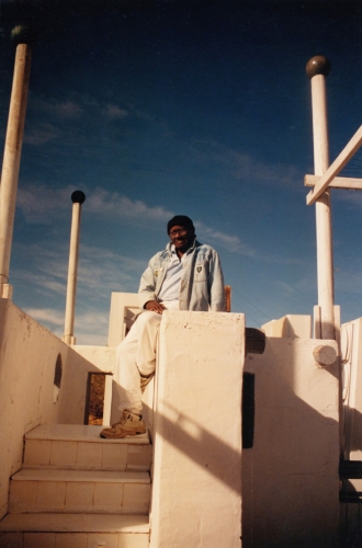 This image depicts the artist, Noah Purifoy, sitting in his artwork, "The White House", which was made from 1990 to 1993.